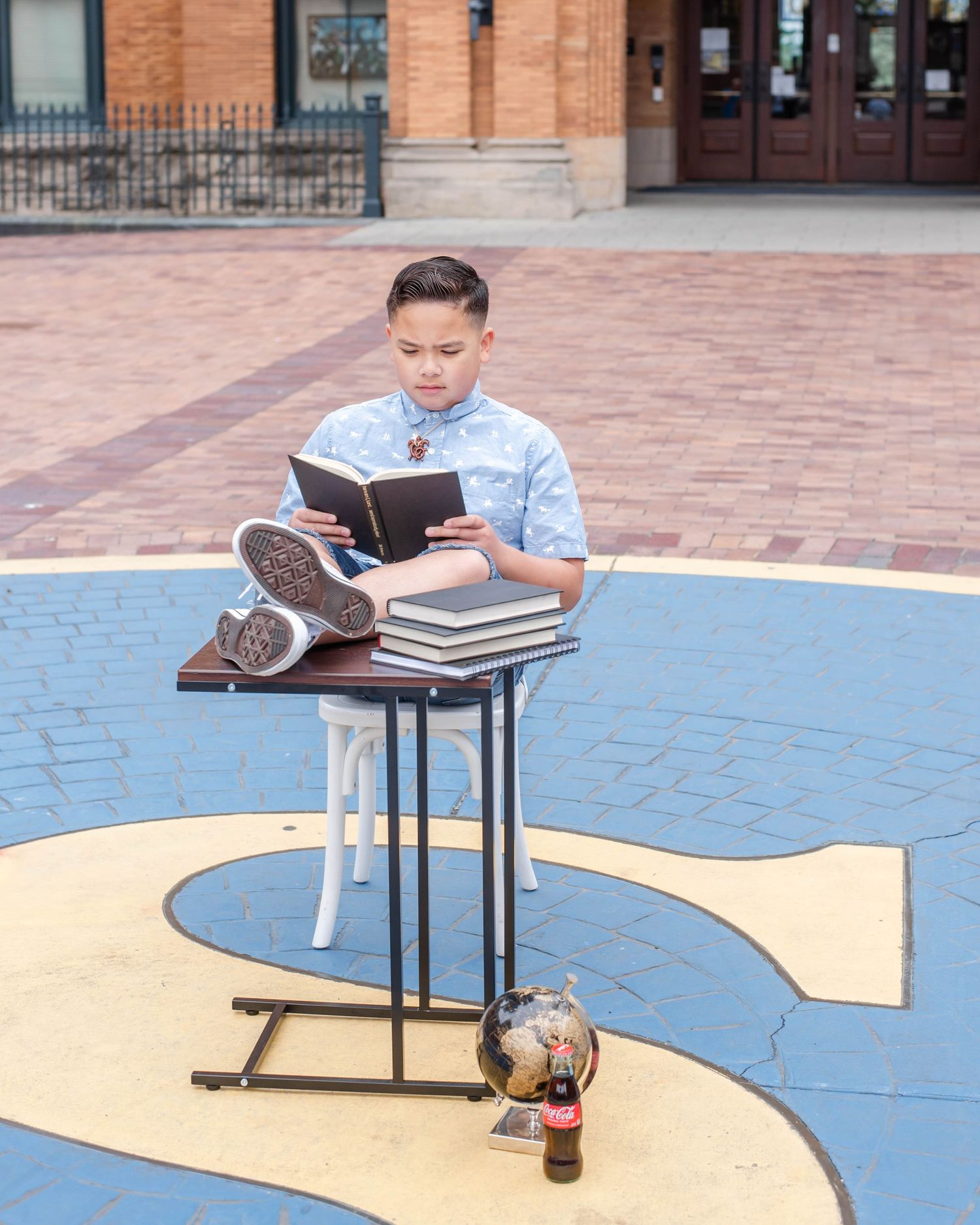 Student reads textbook at small table on blue and yellow paved courtyard.