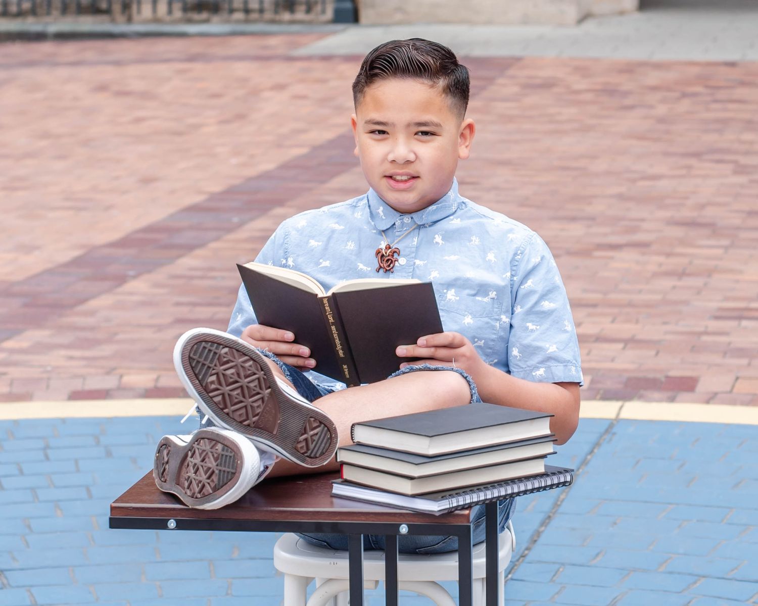 Student reads from book while sitting at small study table outdoors.
