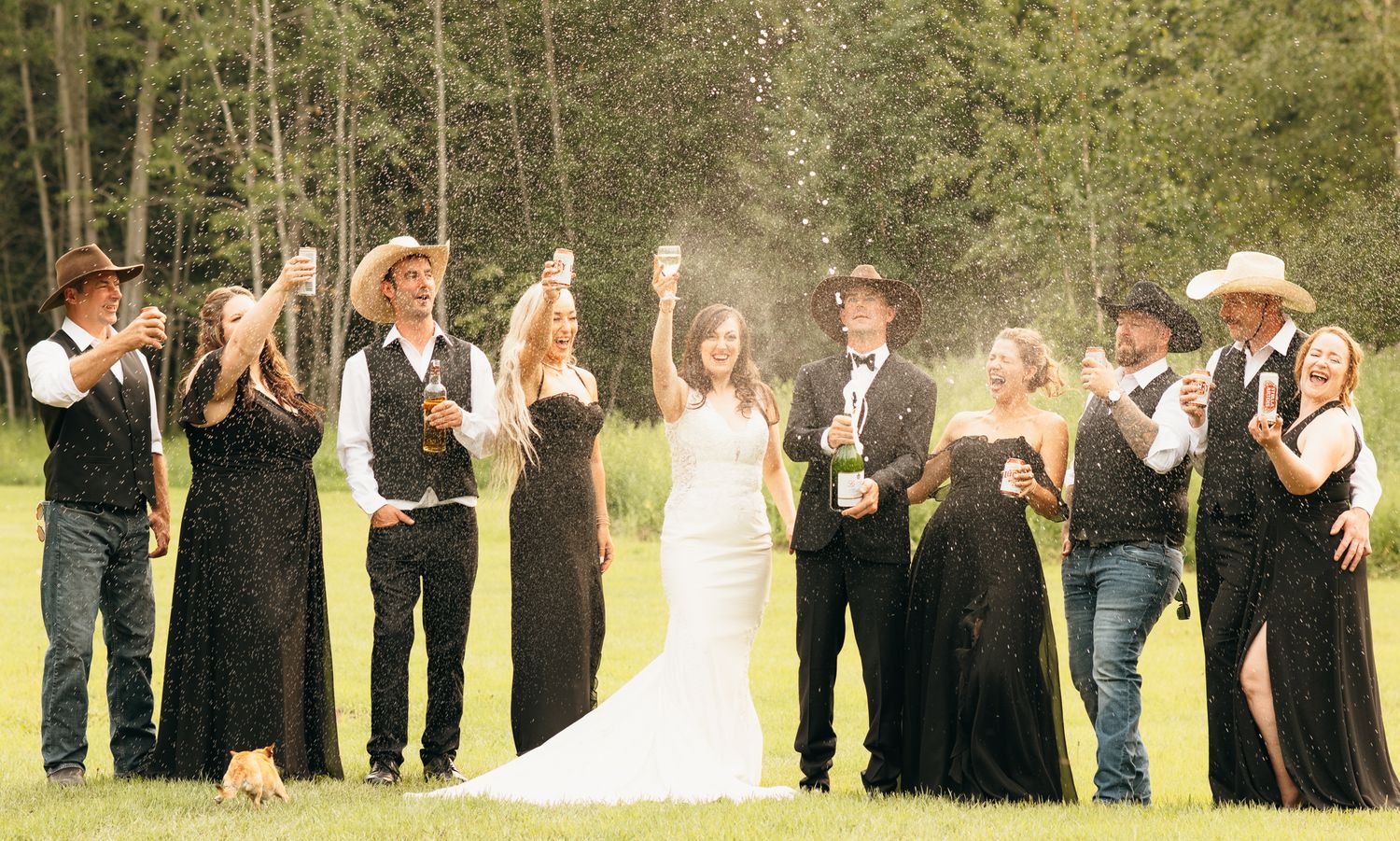 Wedding party celebrating outdoors popping champagne bottles, bridesmaids in black gowns with cowboy-themed groomsmen.
