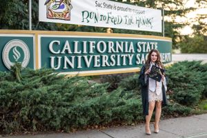 Graduate poses by California State University entrance sign in graduation attire.