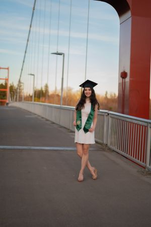 Graduate in white dress and green stole poses on bridge during sunset graduation photoshoot.