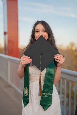 Graduate holding black mortarboard in front of face while wearing white dress and green stole.