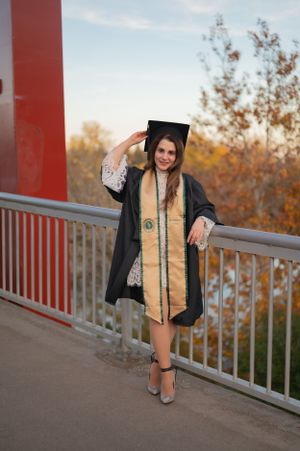 Graduate wearing black cap and gown with gold sash poses on bridge during golden hour.