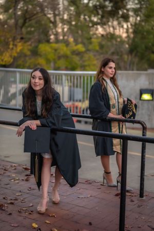 Two graduates in black gowns posing by railing at dusk.