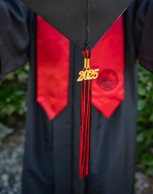 Red graduation tassel with golden 2025 charm hanging from black graduation gown with red accent panels.