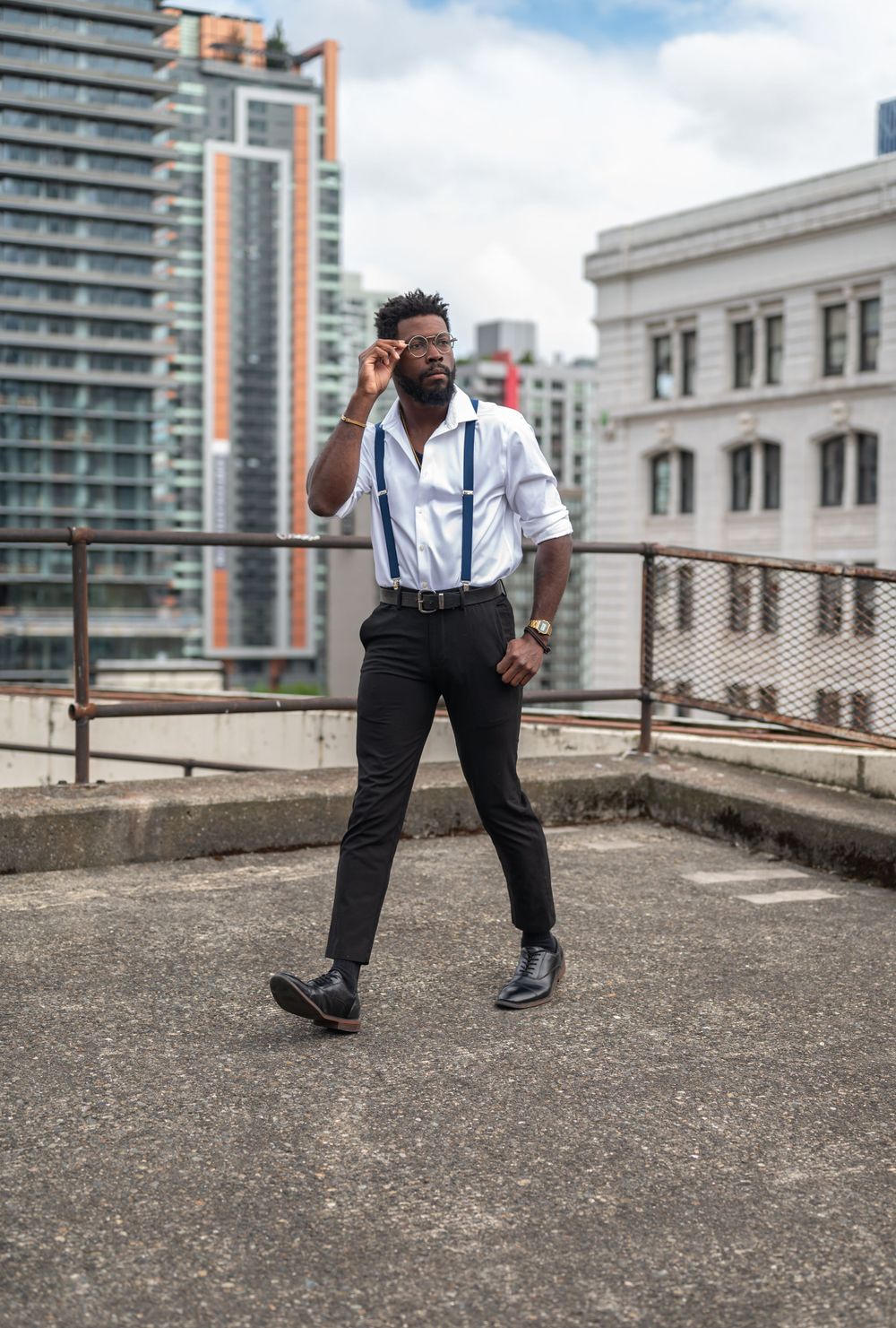 Stylishly dressed person in suspenders and white shirt talking on phone on urban rooftop with city buildings in background.