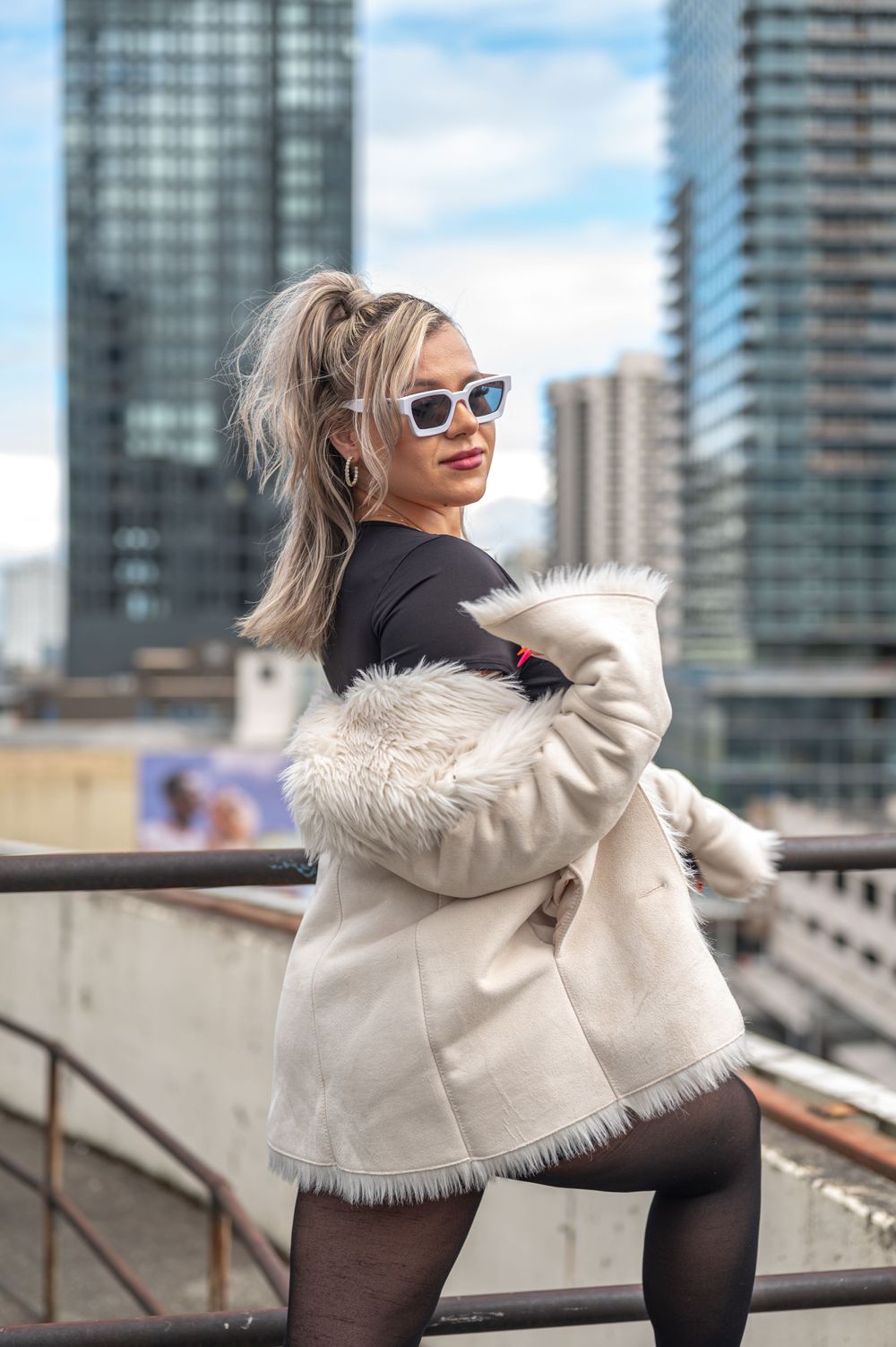Fashion model in furry white jacket and sunglasses poses against cityscape backdrop.