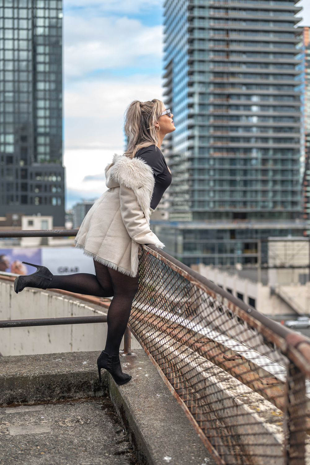 Woman in white fur coat and black outfit poses on rooftop with skyscrapers in background.