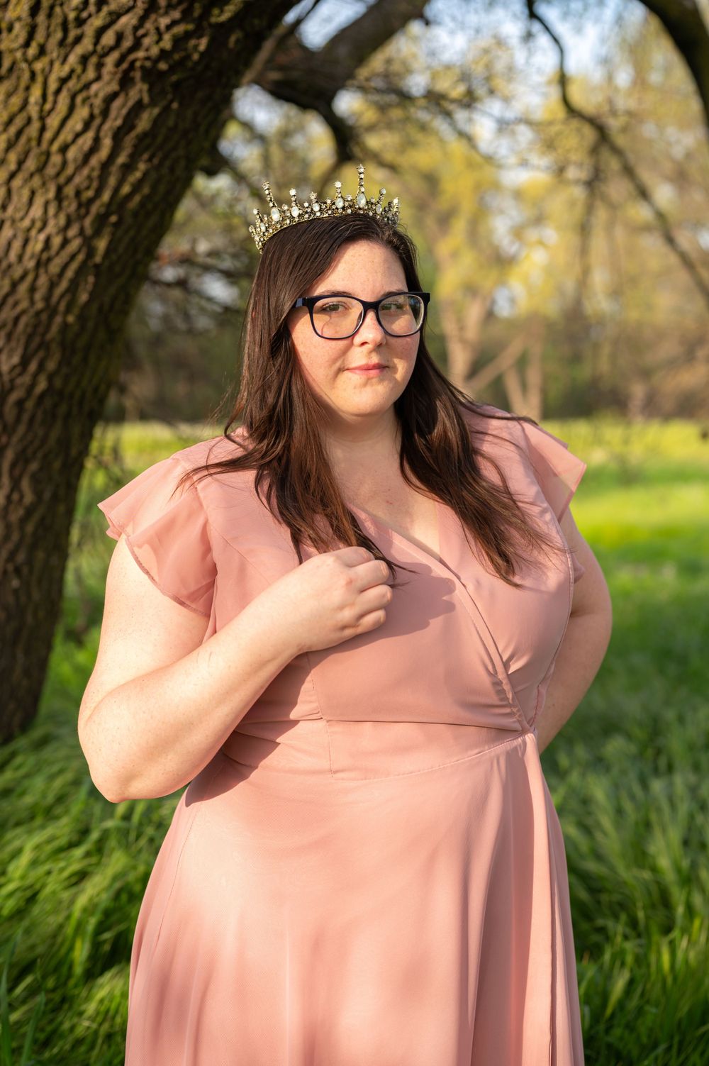 A person in a pink dress and crown poses beneath a tree in a springtime meadow.