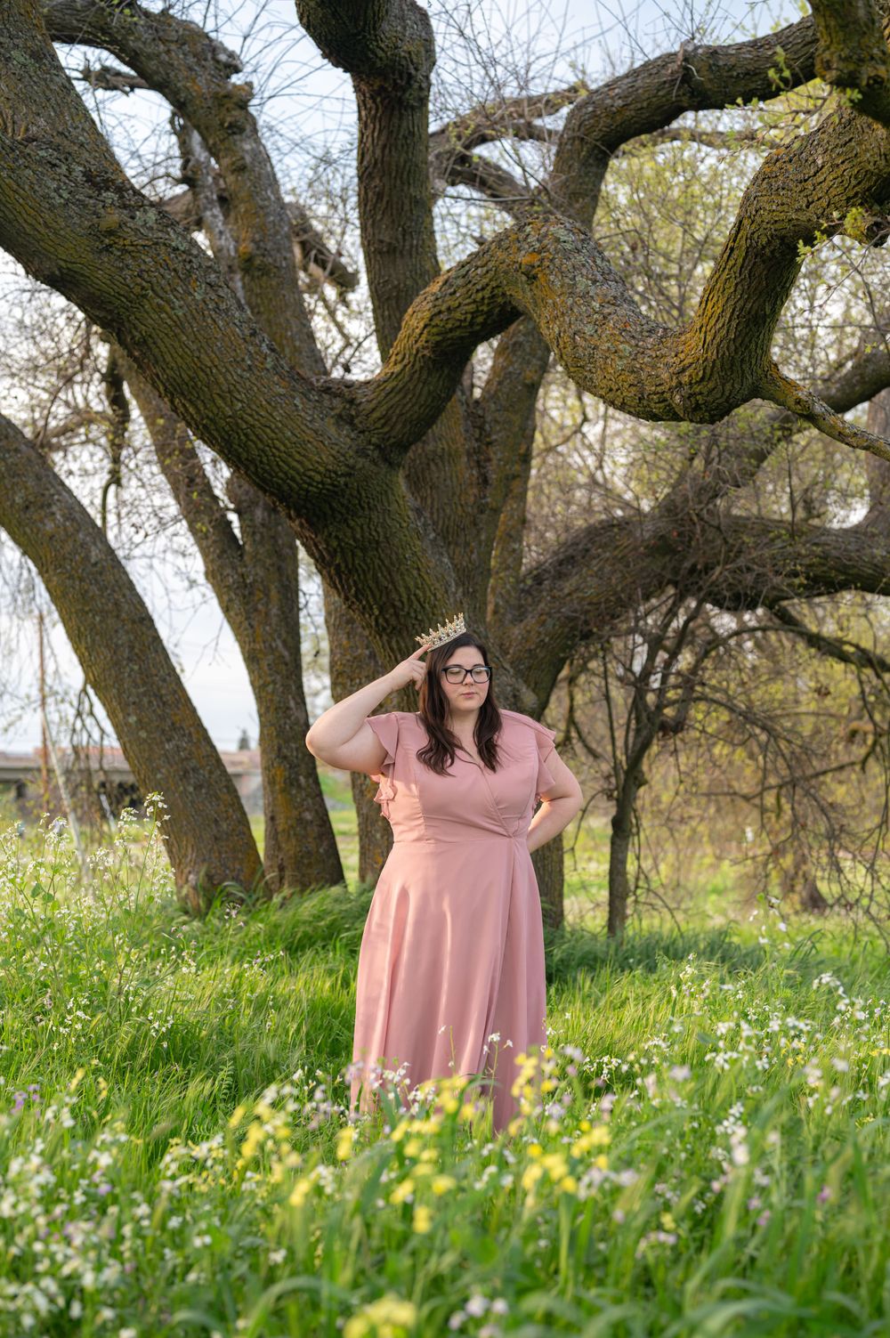 A figure in a pink dress poses in a field of wildflowers beneath a large gnarled tree.