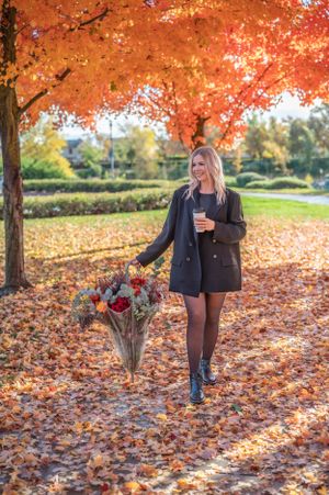 A woman in a black blazer holds flowers under vibrant orange autumn trees.