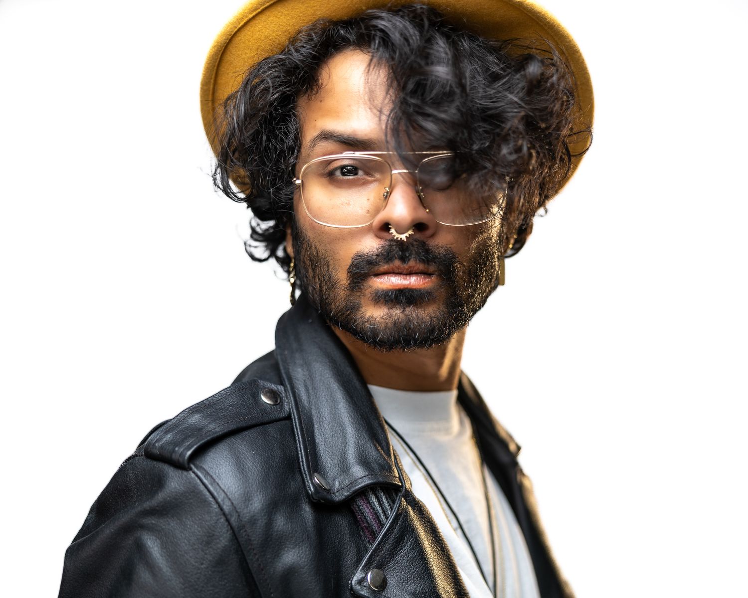 Person wearing leather jacket and brown hat with curly dark hair and wire-framed glasses poses against white background.