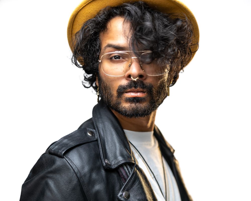 Person wearing leather jacket and brown hat with curly dark hair and wire-framed glasses poses against white background.