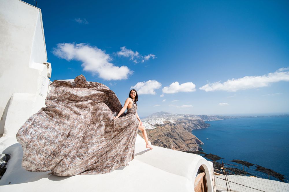 Beige dress billows in wind atop white Santorini rooftop with coastal views.