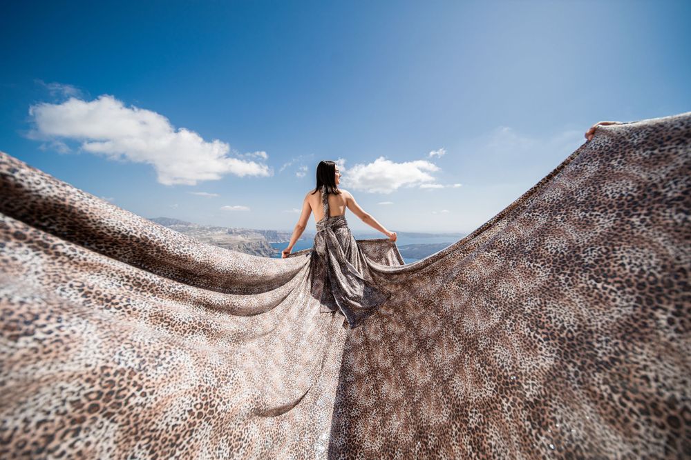 Flowing beige patterned dress billowing dramatically against Mediterranean coastal backdrop in Santorini.