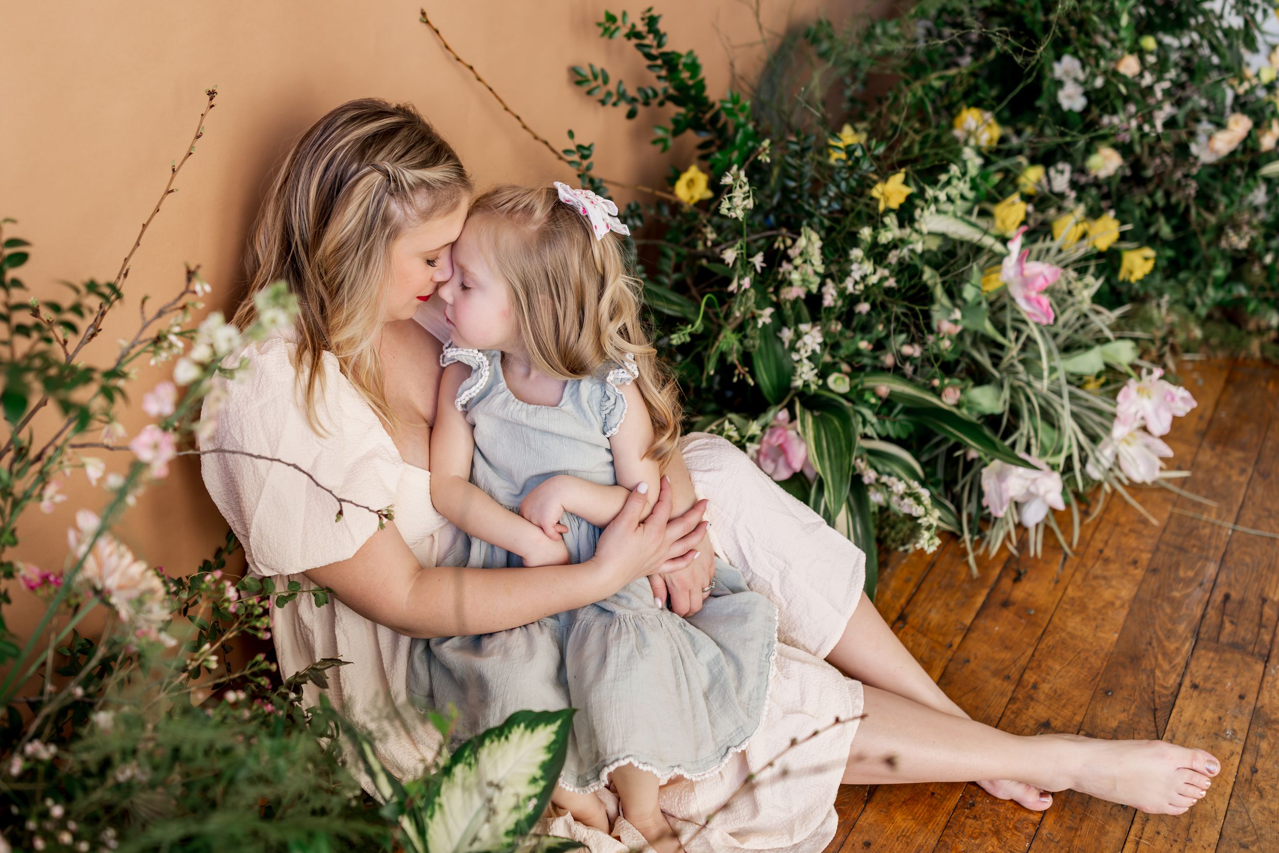 A tender moment shared on wooden floors surrounded by blooming flowers and greenery in soft natural light.
