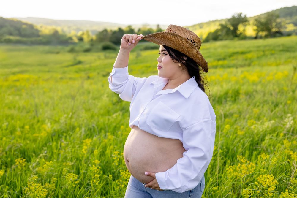 Expectant parent in a white shirt and straw hat stands in a sunny meadow of yellow wildflowers.