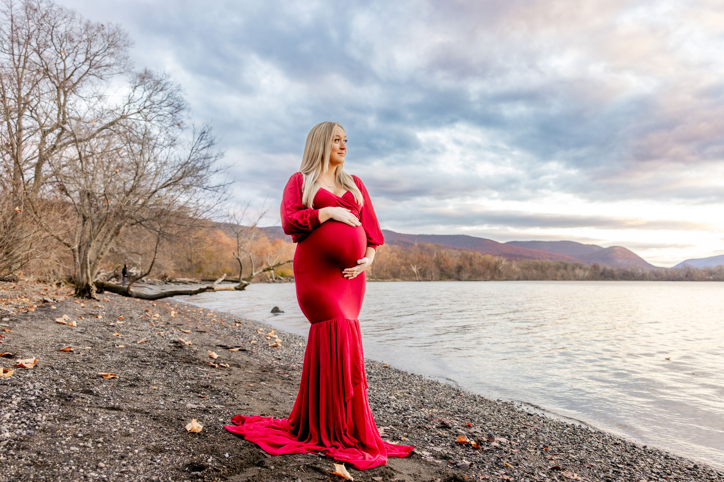 A figure in a flowing red dress stands on a rocky shoreline at sunset with mountains and a lake in the background.
