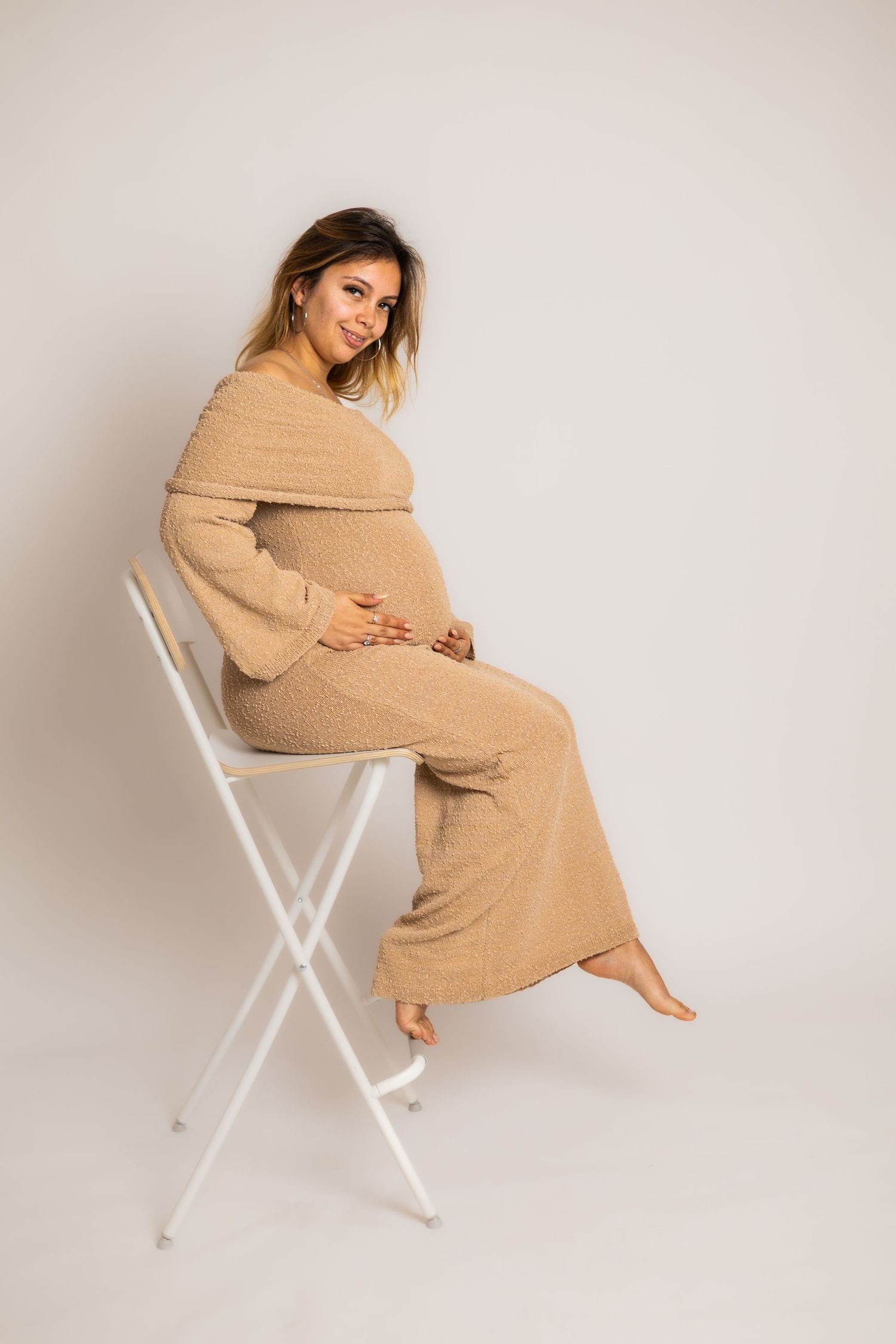 Cozy maternity photos featuring woman in beige sweater dress posing on white chair.