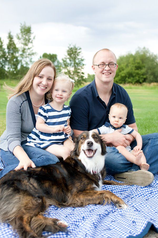 A happy family enjoys a picnic on a checkered blanket with their Australian Shepherd dog in a green field.