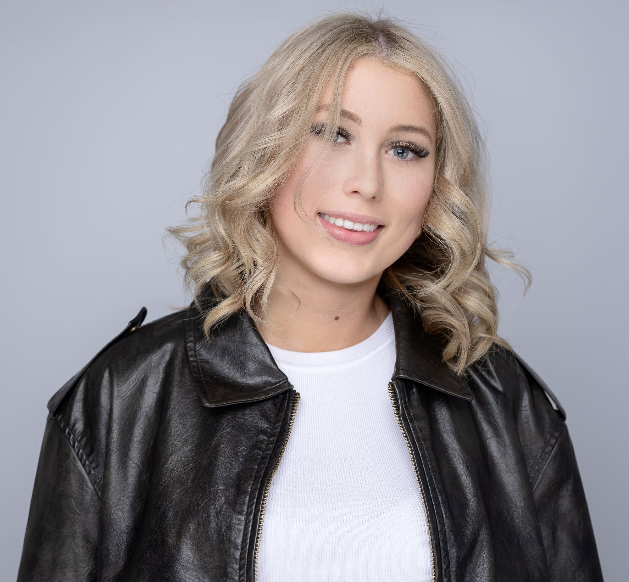 Professional headshot with wavy blonde hair wearing leather jacket against grey backdrop.