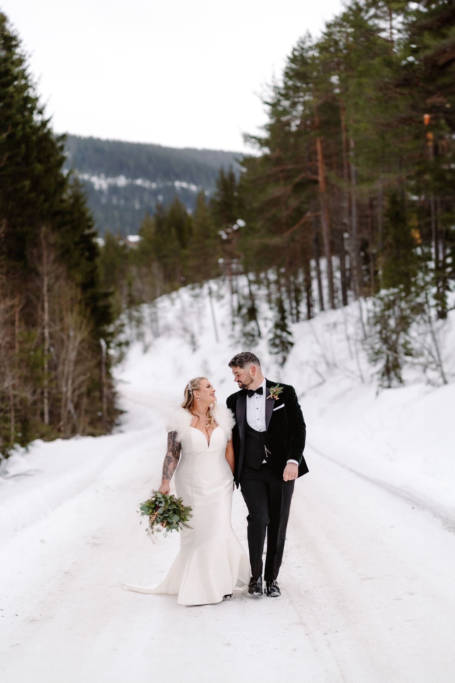Winter wedding couple walking through a snow-covered forest path surrounded by tall pine trees.