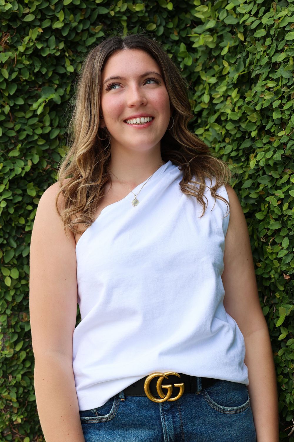 Casual summer style with white one-shoulder top, Gucci belt and jeans against lush green hedge.