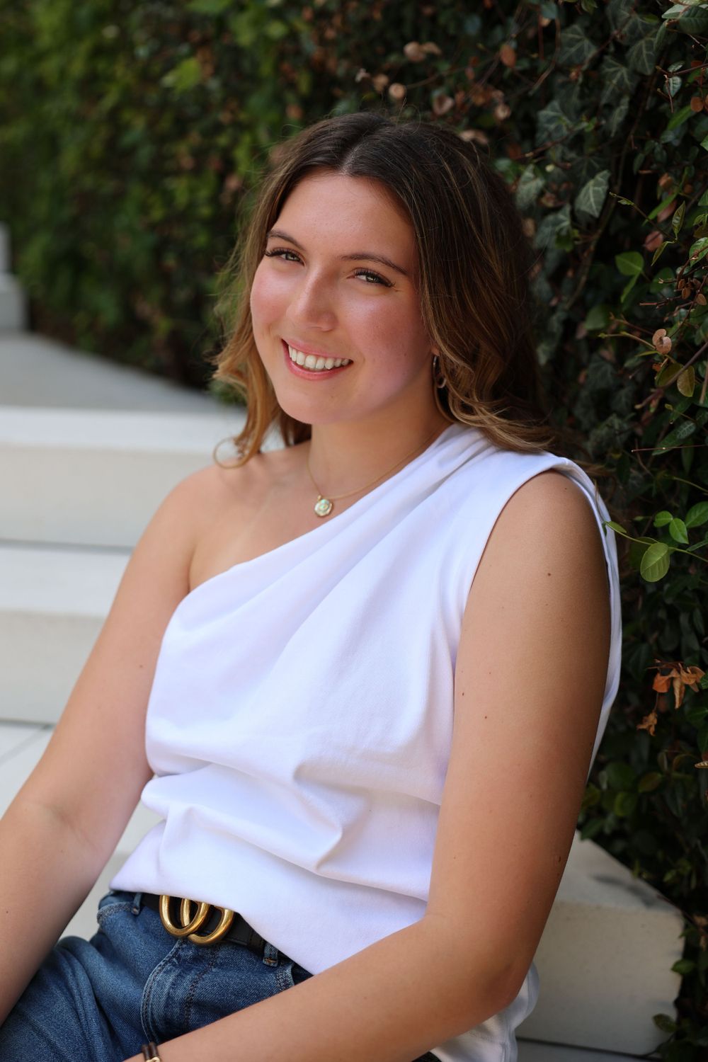 Summer fashion look with asymmetrical white top and designer belt against greenery backdrop.