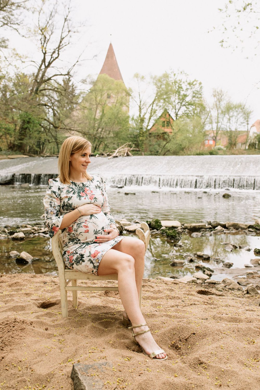 Woman in floral dress sitting by waterfall and church spire in scenic outdoor setting.