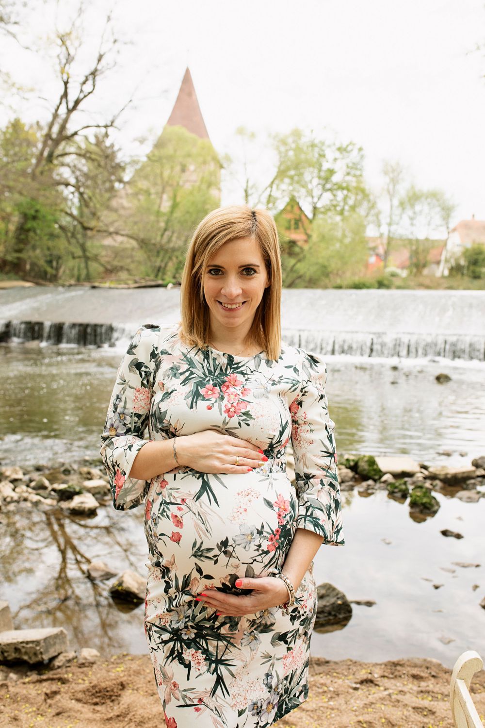 A floral maternity dress is showcased against a scenic waterfall backdrop in a springtime outdoor photo session.