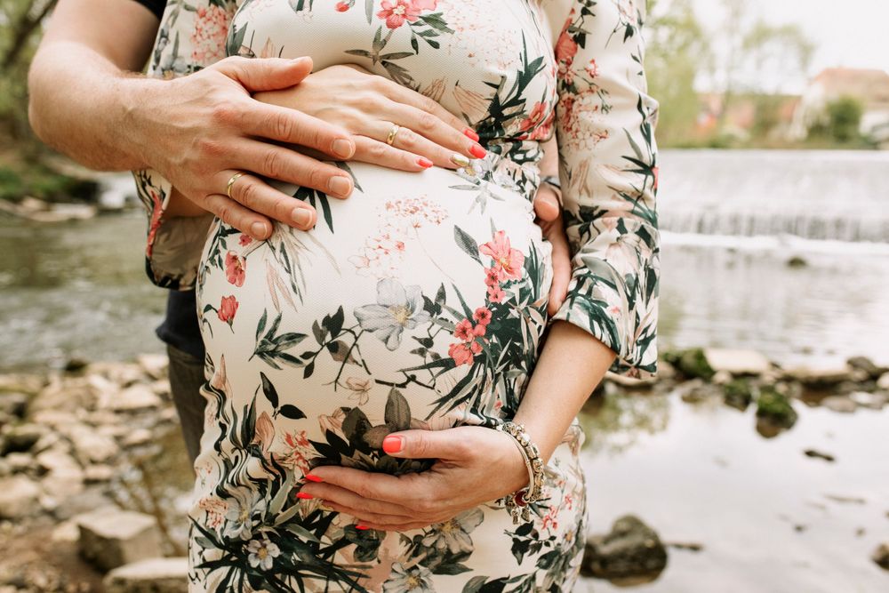 Gentle hands cradle a pregnant belly wearing a white floral dress by a waterfall.