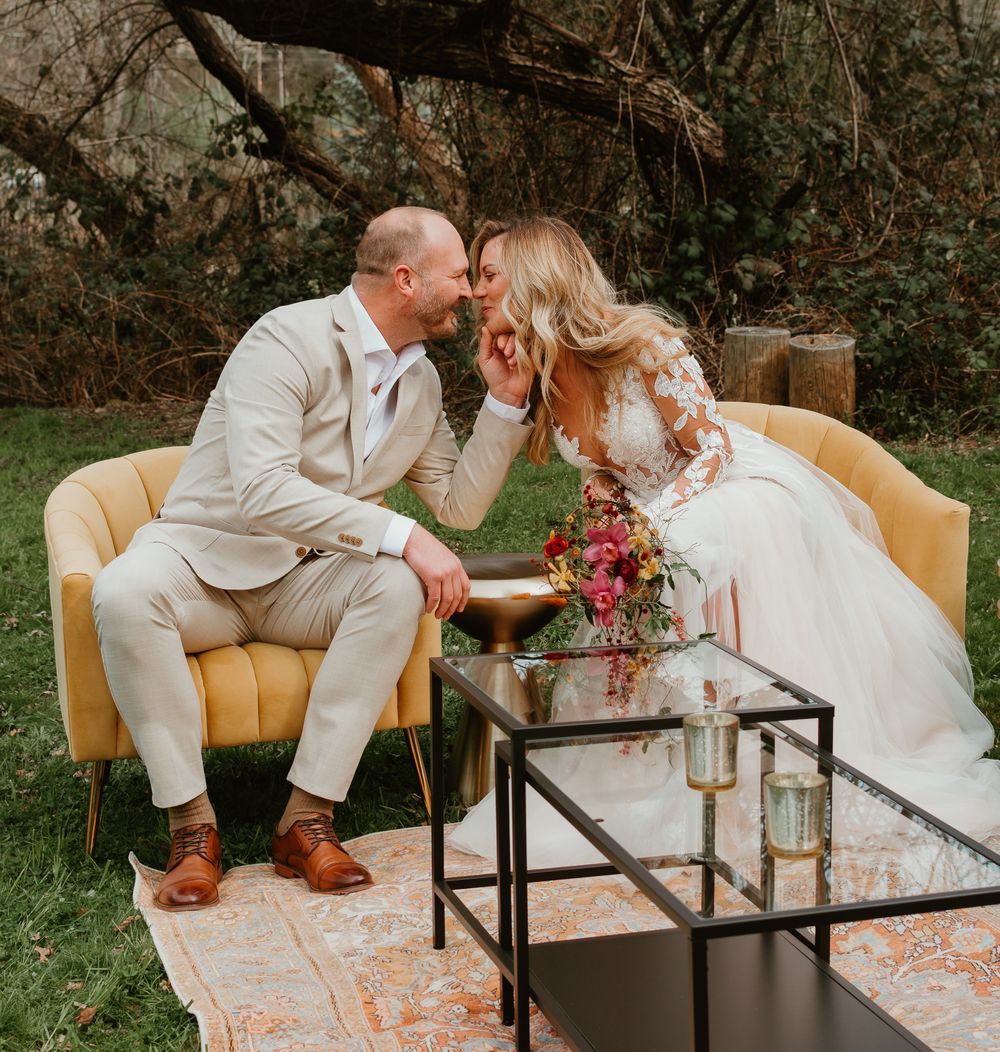 Wedding couple seated on vintage yellow chairs with glass coffee table outdoors.