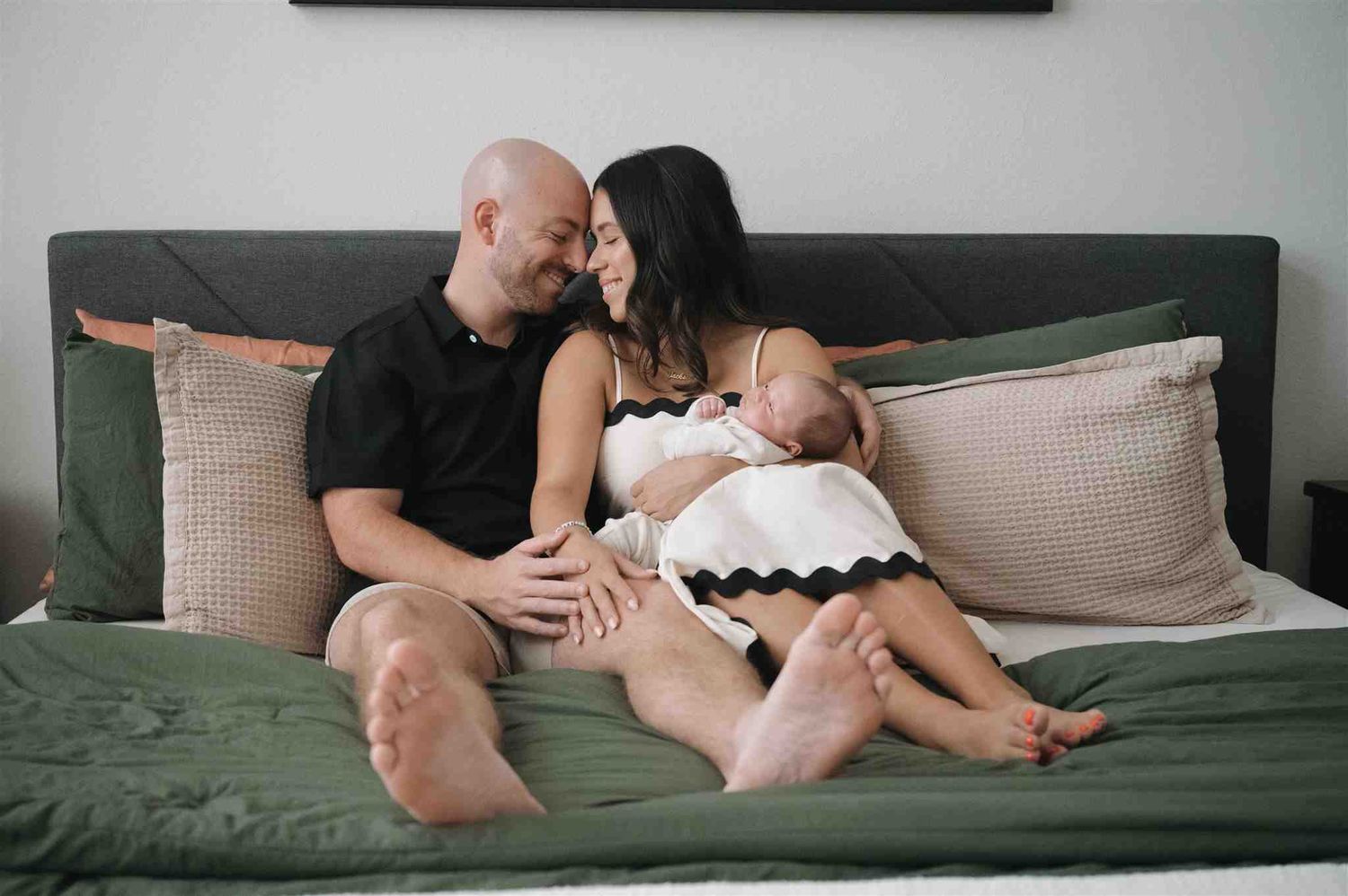 A couple sits on a green couch with throw pillows, sharing a tender moment while holding a baby.