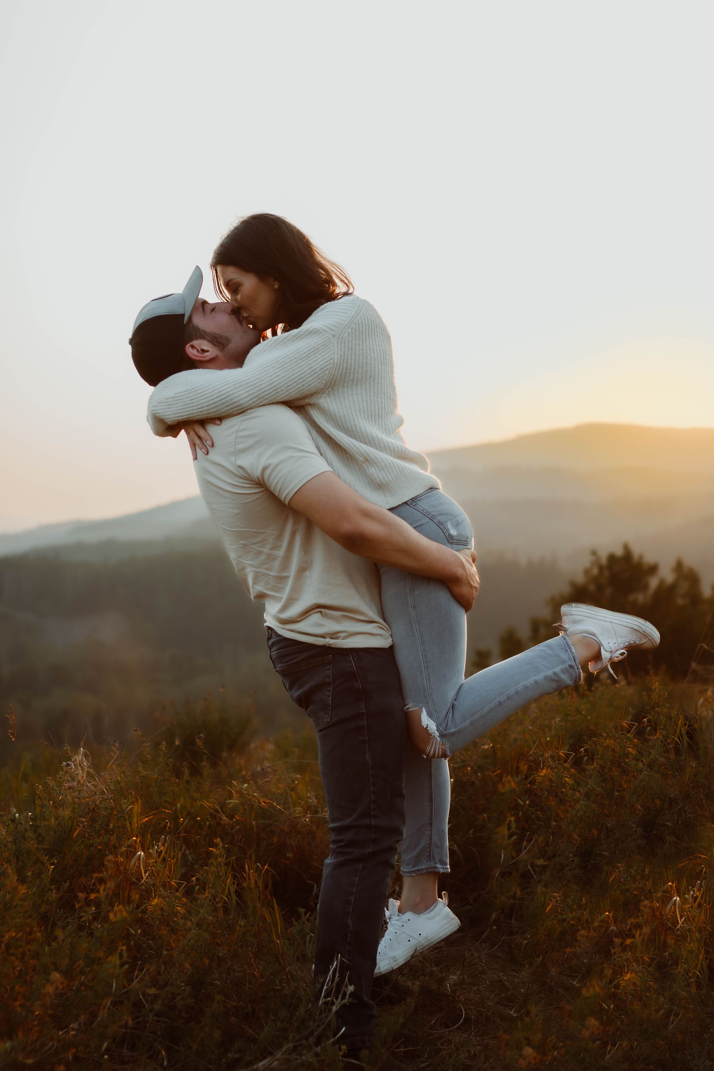 Couple embraces during sunset in mountainous landscape while wearing casual clothing.
