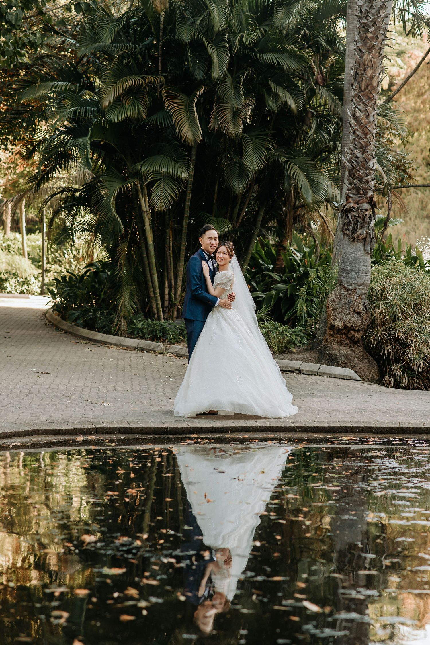 A romantic wedding portrait by a reflective pond surrounded by tropical palm trees.