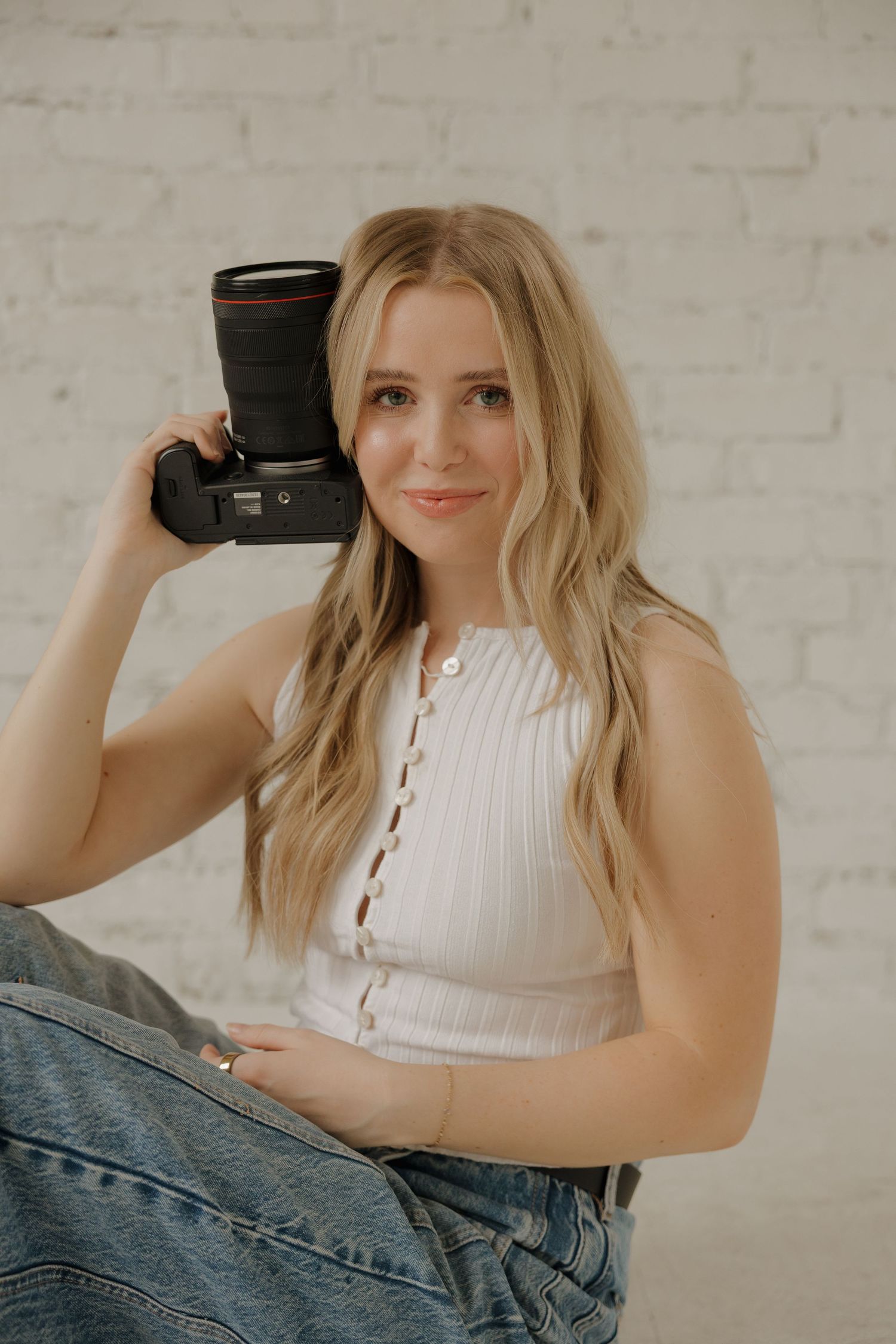 Smiling blonde female photographer holding a camera beside her face against a white brick wall.