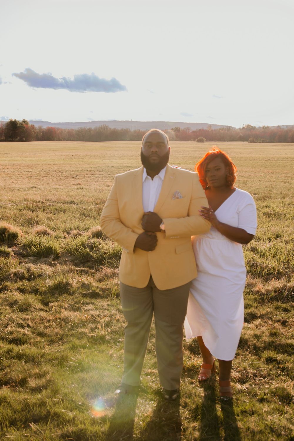 Elegant couple in yellow suit and white dress pose together in sunlit field.