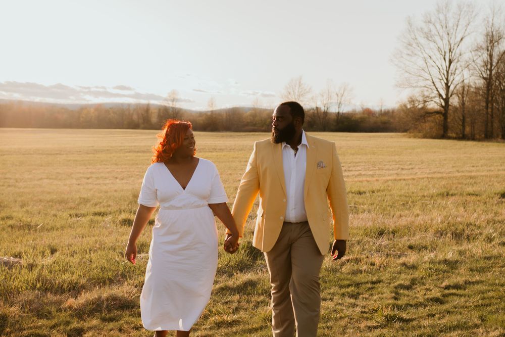 A couple holding hands walks through a golden field at sunset with mountains in the background.