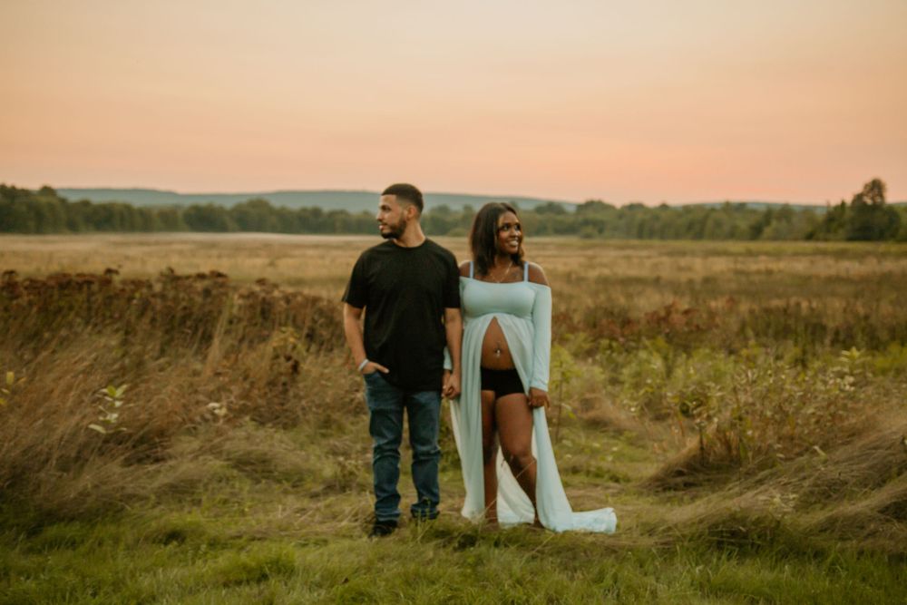 Sunset maternity session with couple standing together in grassy field.
