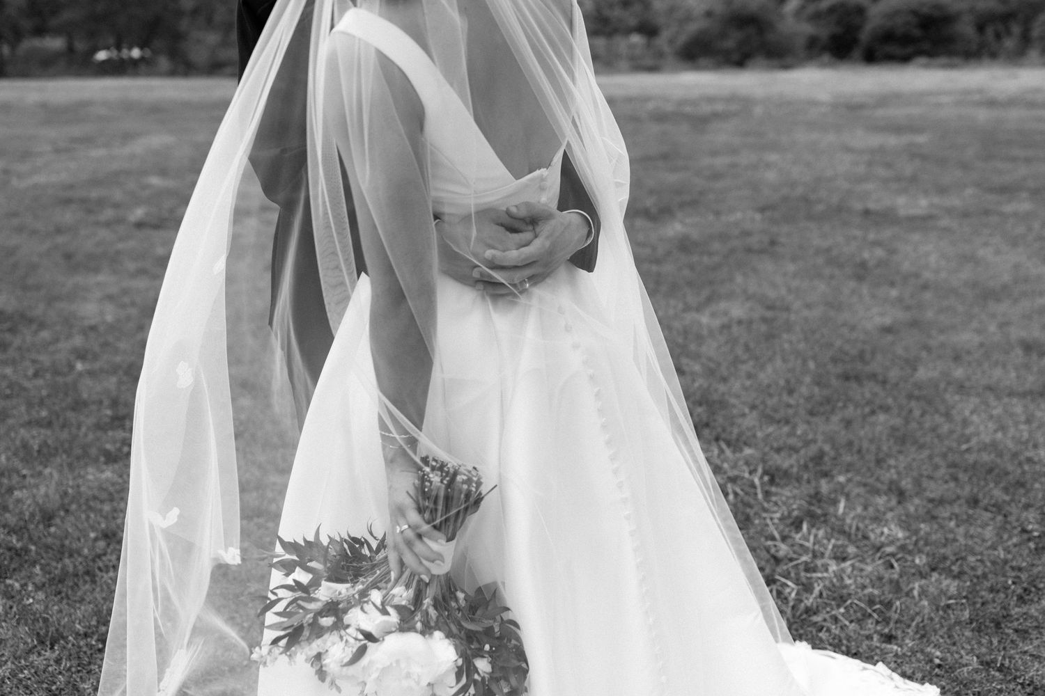 A flowing white wedding dress and veil photographed from behind in a garden setting.