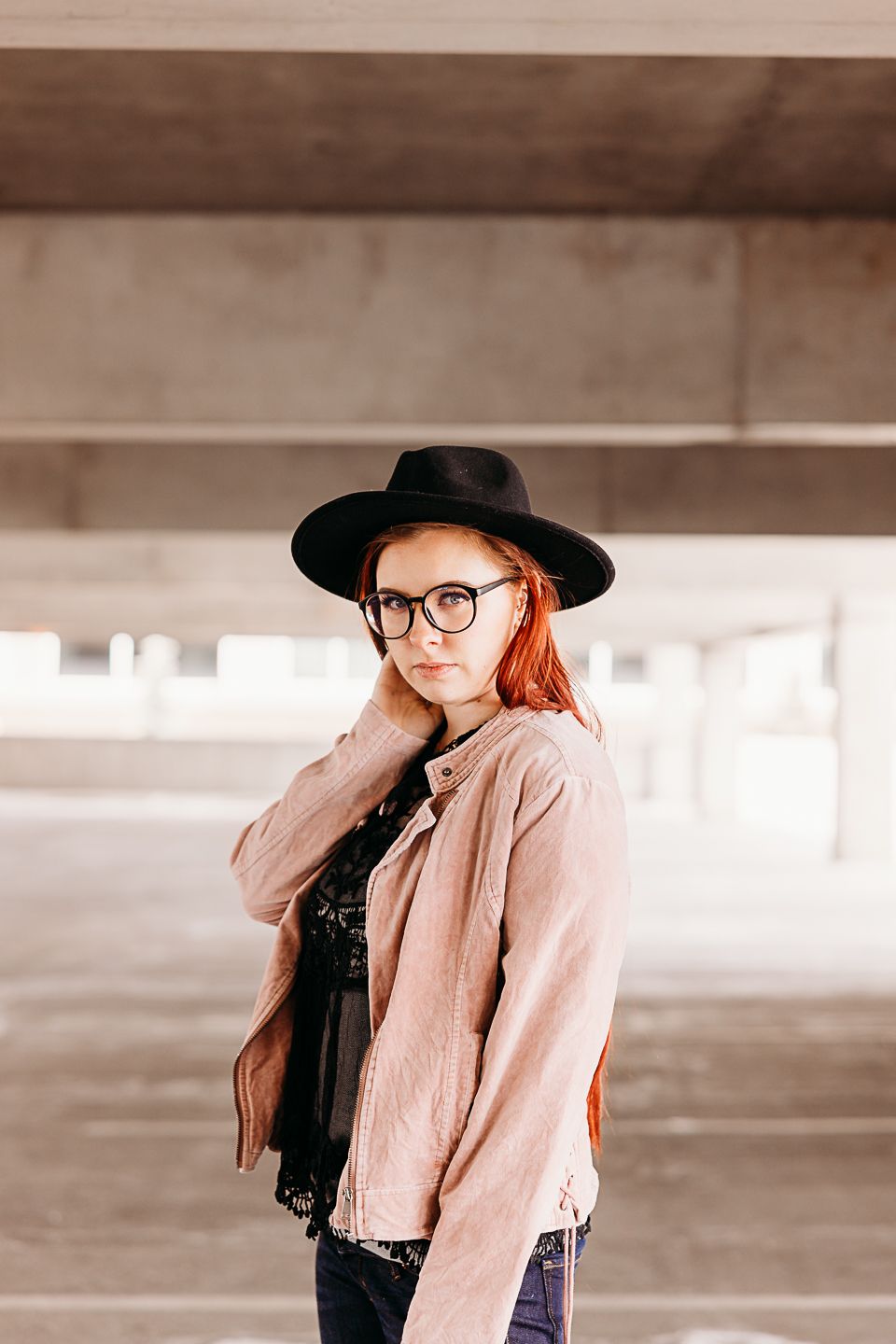 Person wearing black hat and pink jacket poses in minimalist concrete parking structure.