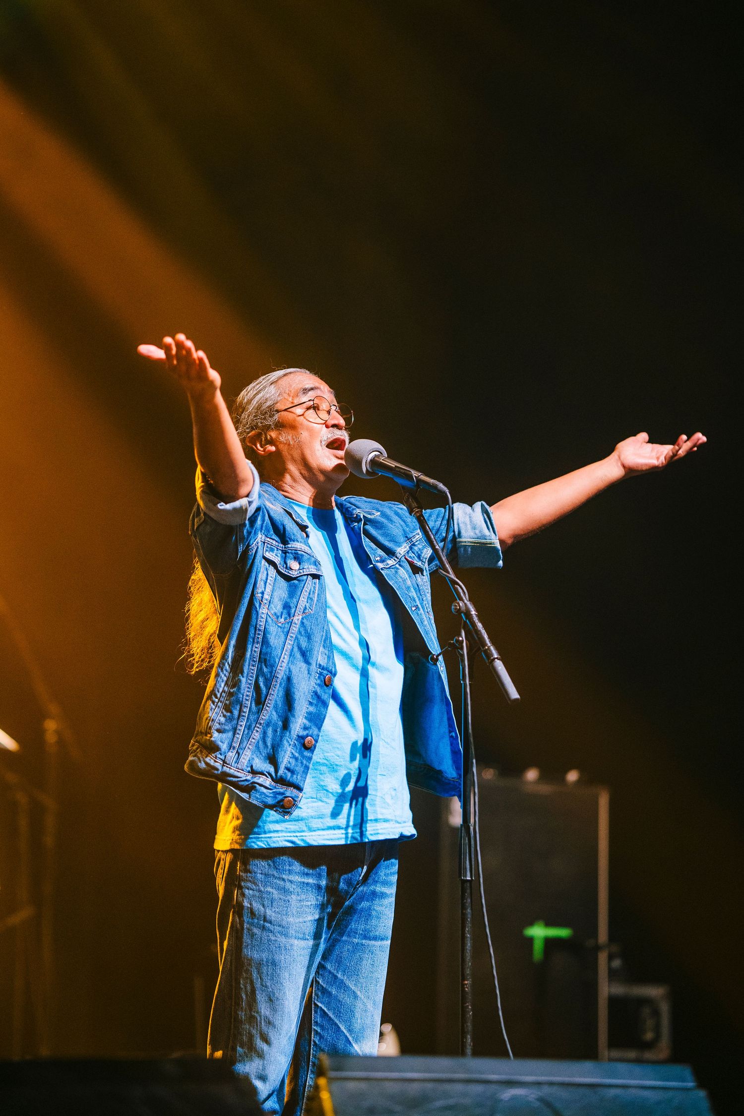 Performer raises arms wide while singing passionately on stage with orange lighting effects.