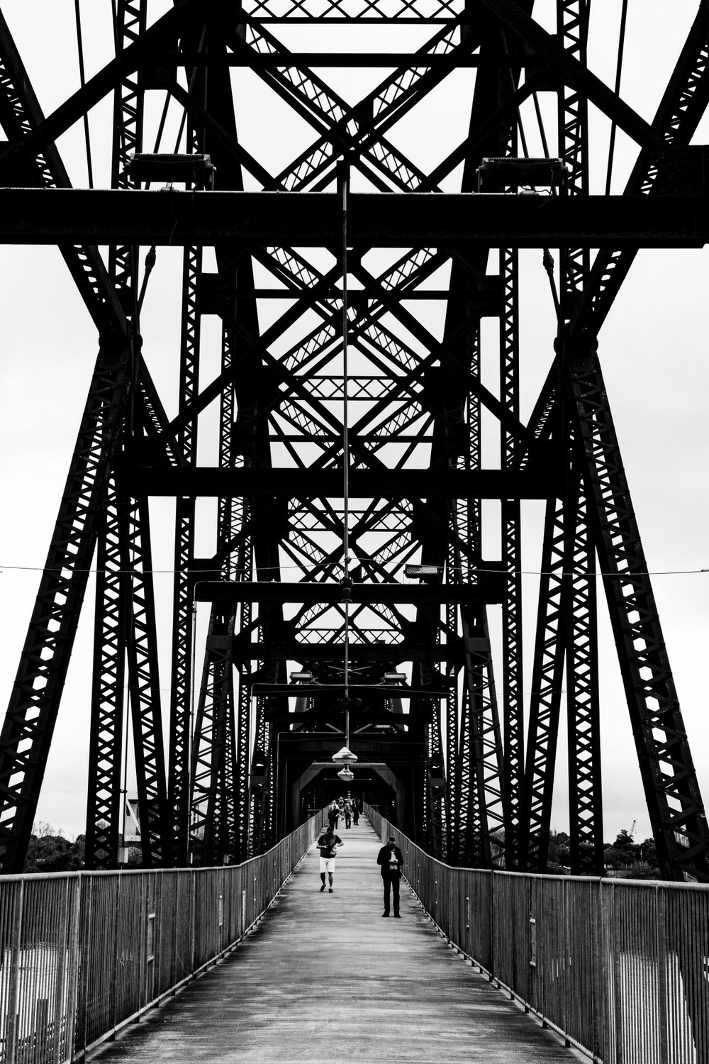 Black and white perspective of steel bridge walkway with geometric patterns and pedestrians.