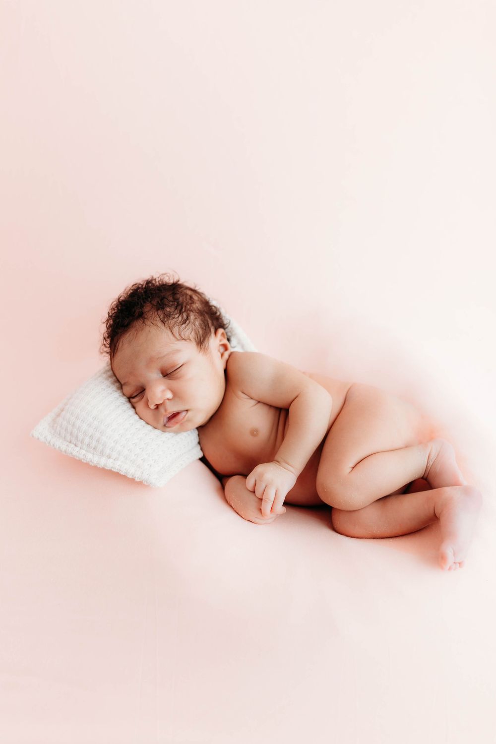 Sleeping newborn posed on white pillow against soft pink background.