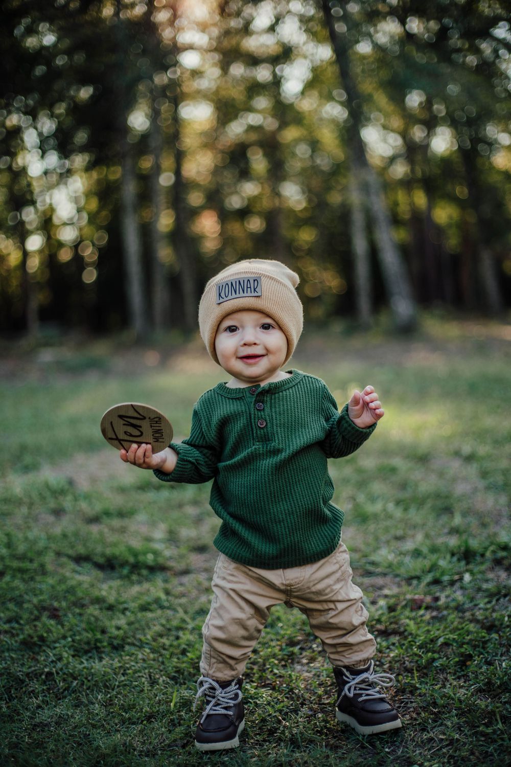 Toddler in green sweater and beanie hat playing outdoors with autumn trees in background.