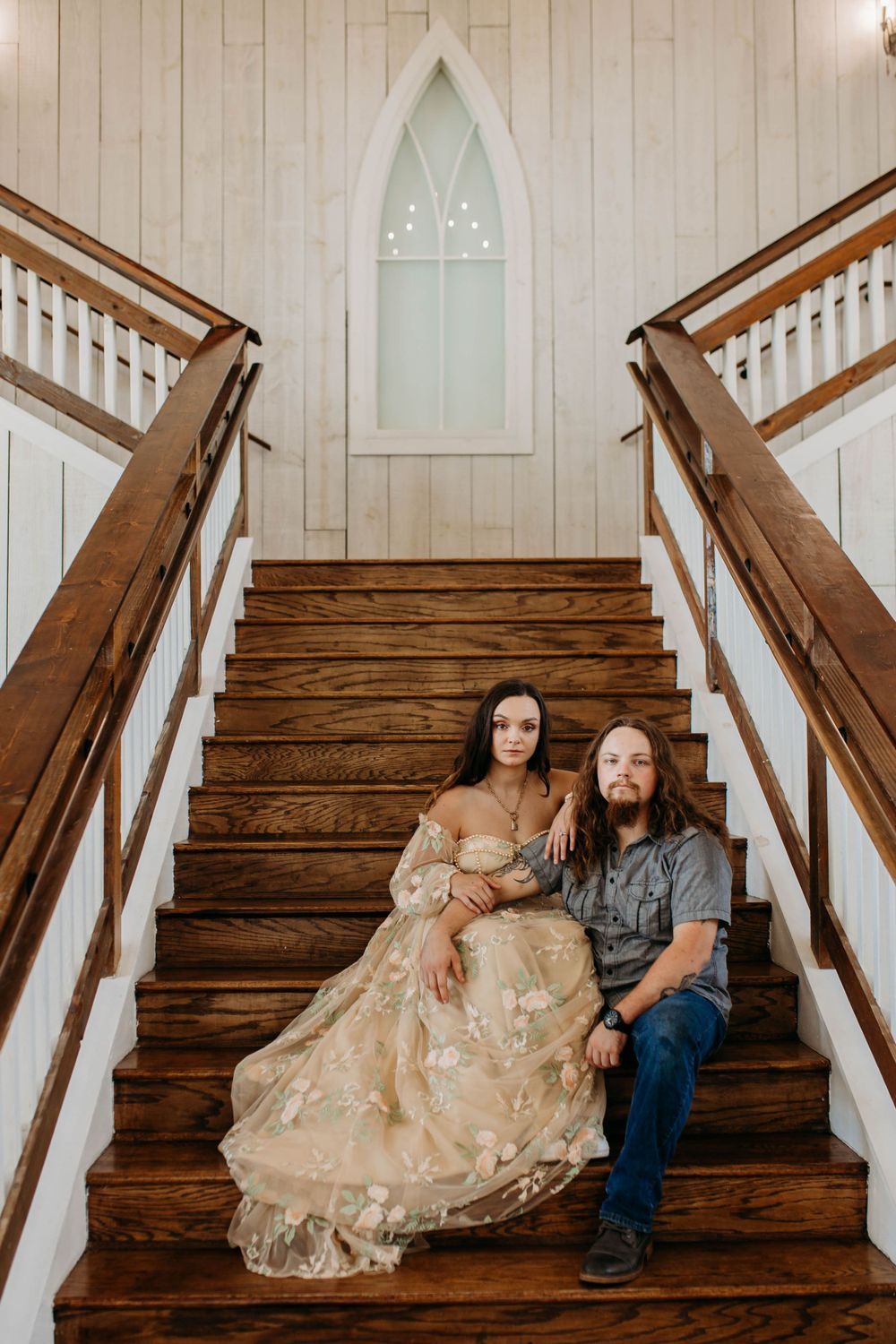 Couple in vintage-style clothing sits together on wooden church steps beneath arched window.