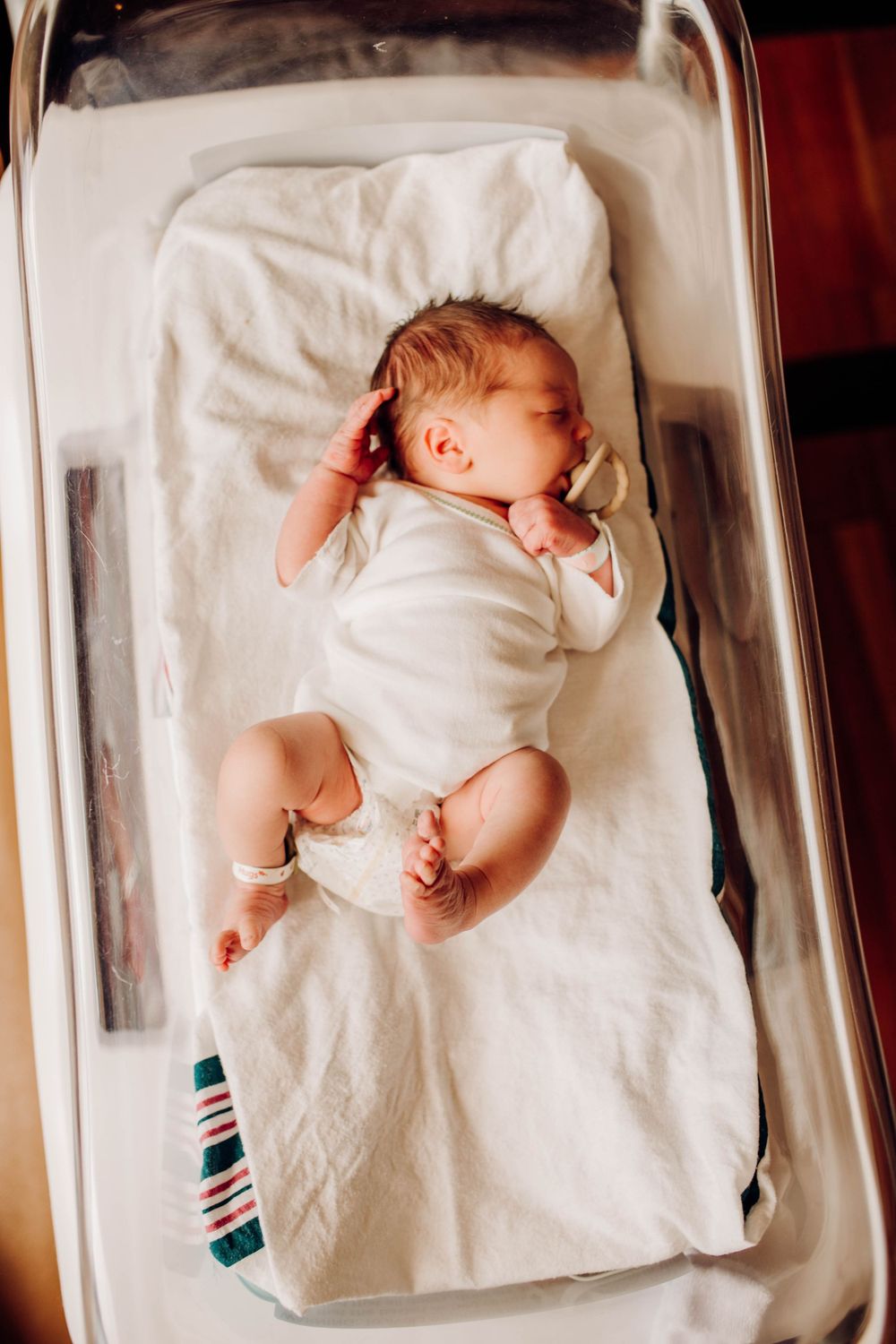 Newborn sleeping peacefully in white hospital bassinet wearing white onesie.