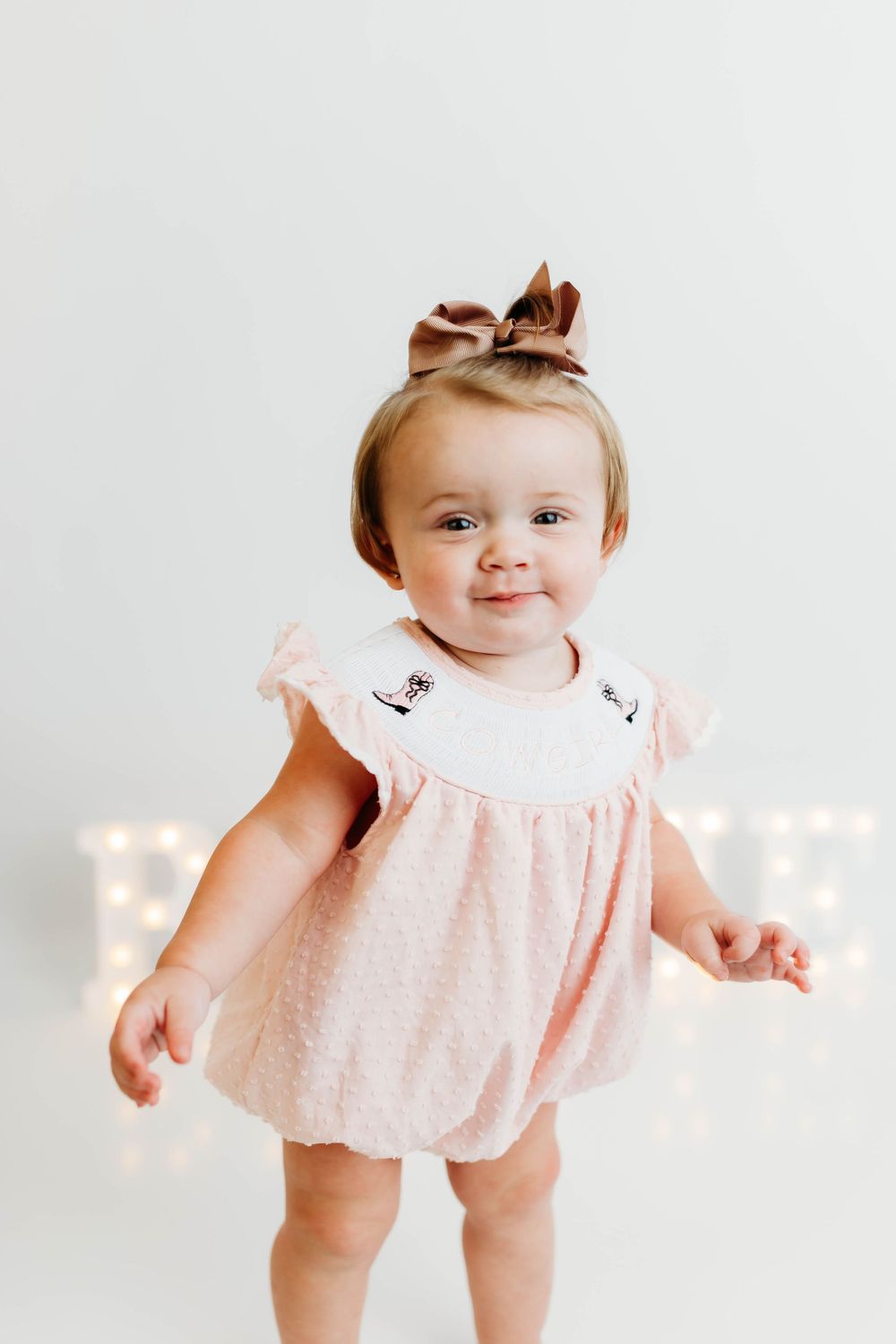 Baby wearing pink dress with bow headband smiles while standing in front of white backdrop with twinkle lights.