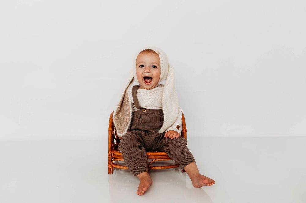 Baby in brown overalls laughing while seated in an orange wooden chair against white background.