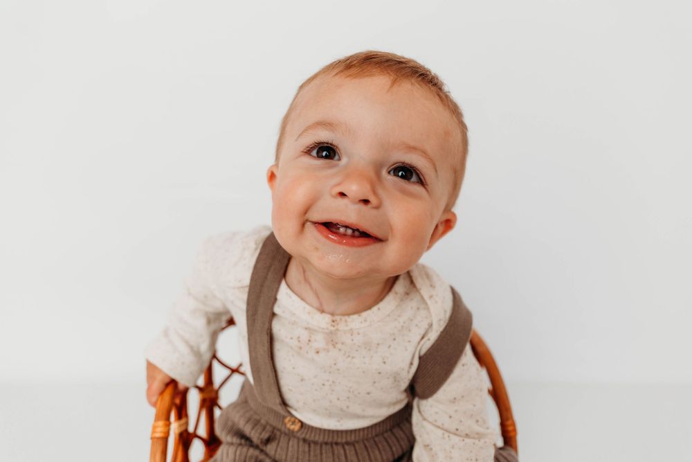 A cheerful baby wearing brown overalls and white shirt sitting in a vintage wooden chair.